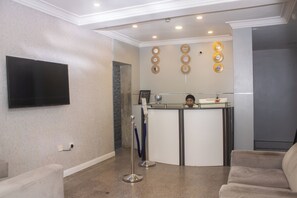 A hotel reception area with a flat-screen TV mounted on the wall, a reception desk with a glass top, and a seating area with a sofa. - Mayors & Diplomat Suites Opebi (Lagos)