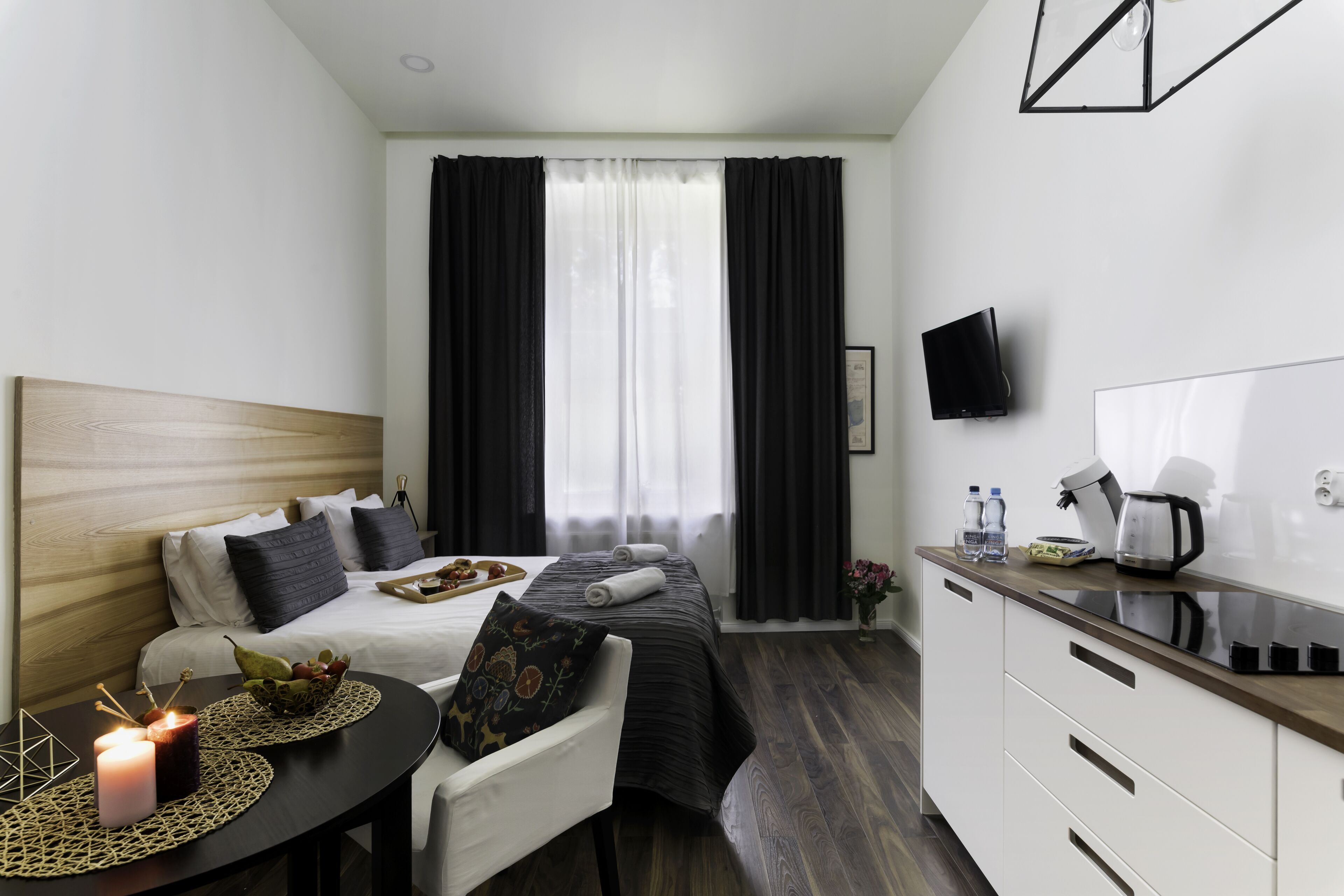 A modern hotel room with a bed, bedside table, and a kitchenette.