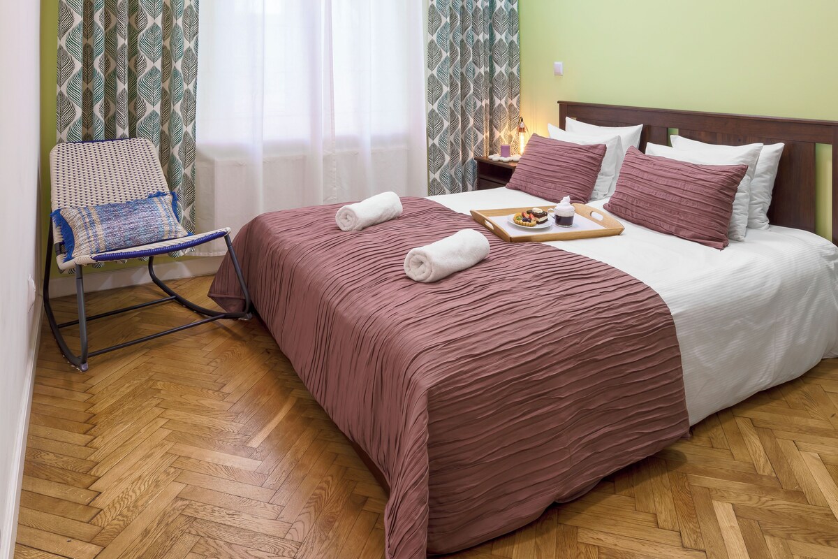 Una cama bien tendida con una colcha marrón y almohadas, una bandeja con comida y bebida, y una silla con un cojín estampado.