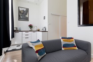 Ein modernes Wohnzimmer mit einem grauen Sofa, bunten Kissen, einer Küchenzeile mit weißen Schränken und einem gerahmten Bild an der Wand.