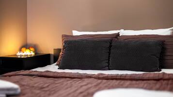 Una cama bien hecha con una manta marrón, dos almohadas oscuras y una almohada blanca. Hay una mesita de noche con una lámpara pequeña y velas.