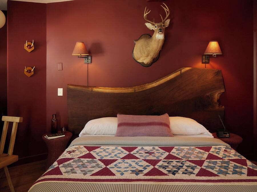 A bedroom with a wooden headboard, a quilt, two wall-mounted lamps, and a mounted deer head.