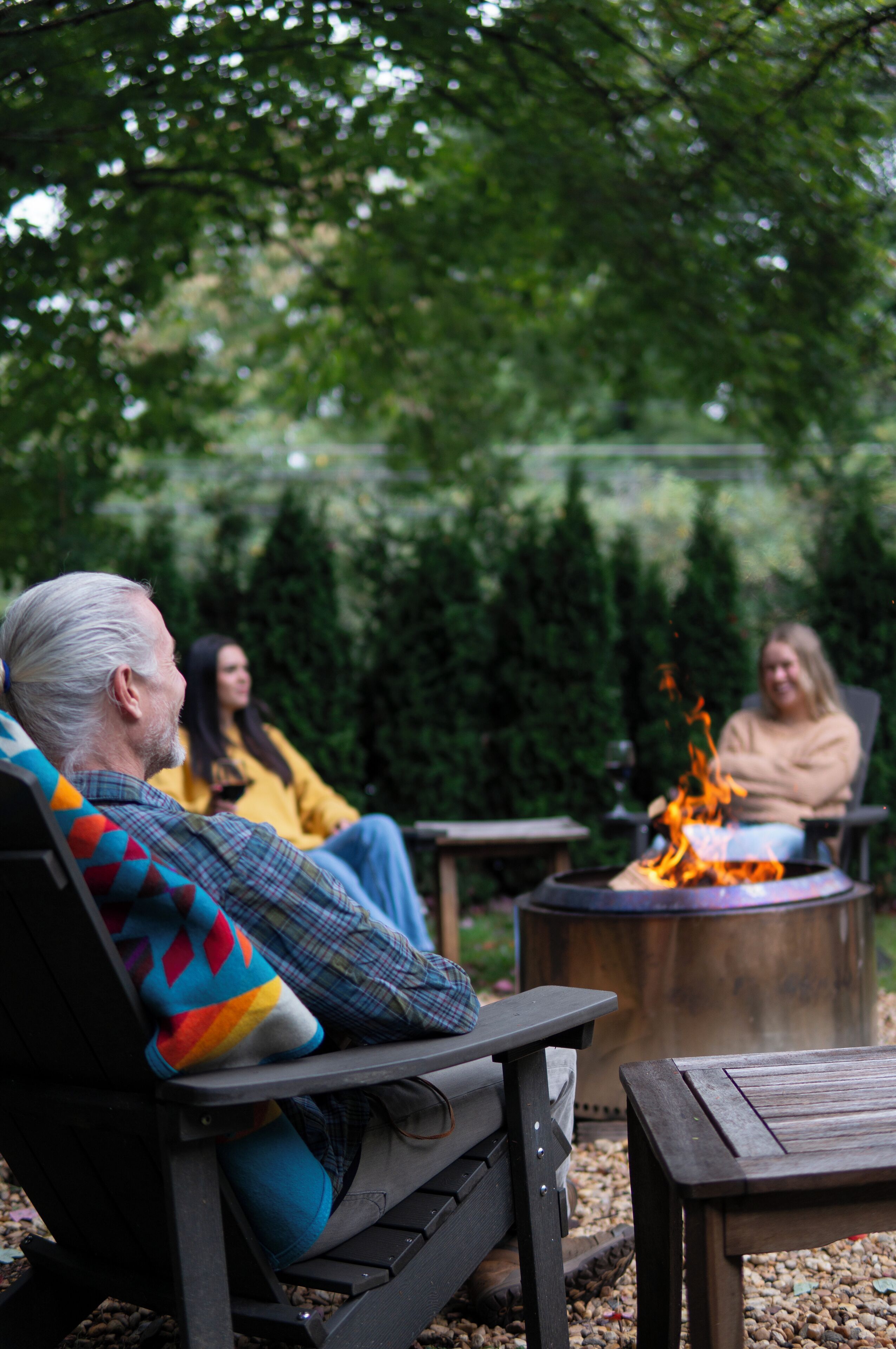 Drie personen zitten op buitenmeubilair rond een vuurkuil in een tuin.