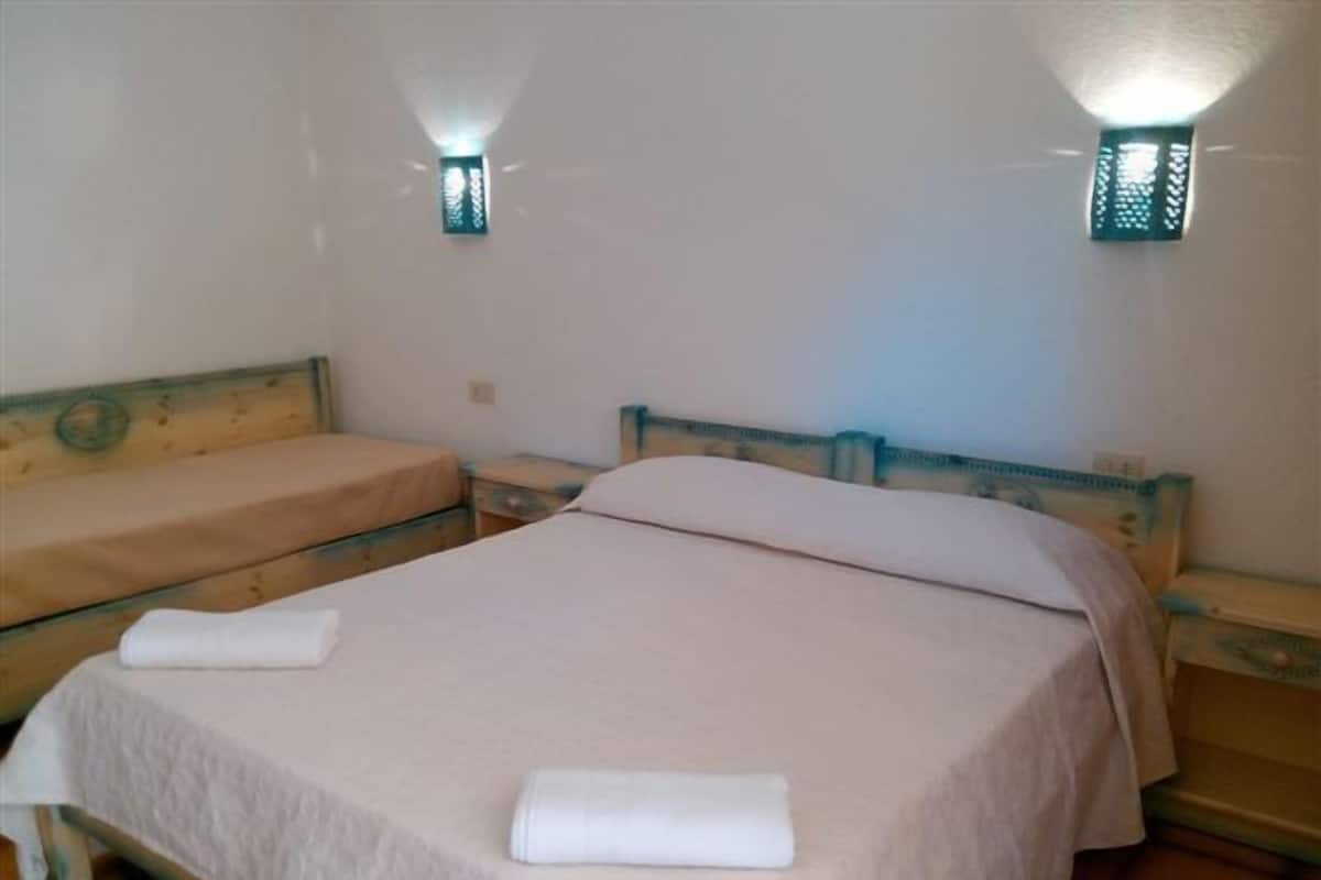 A room with two single beds, each with a wooden headboard and a white bedspread. There are two wall-mounted light fixtures on either side of the room.