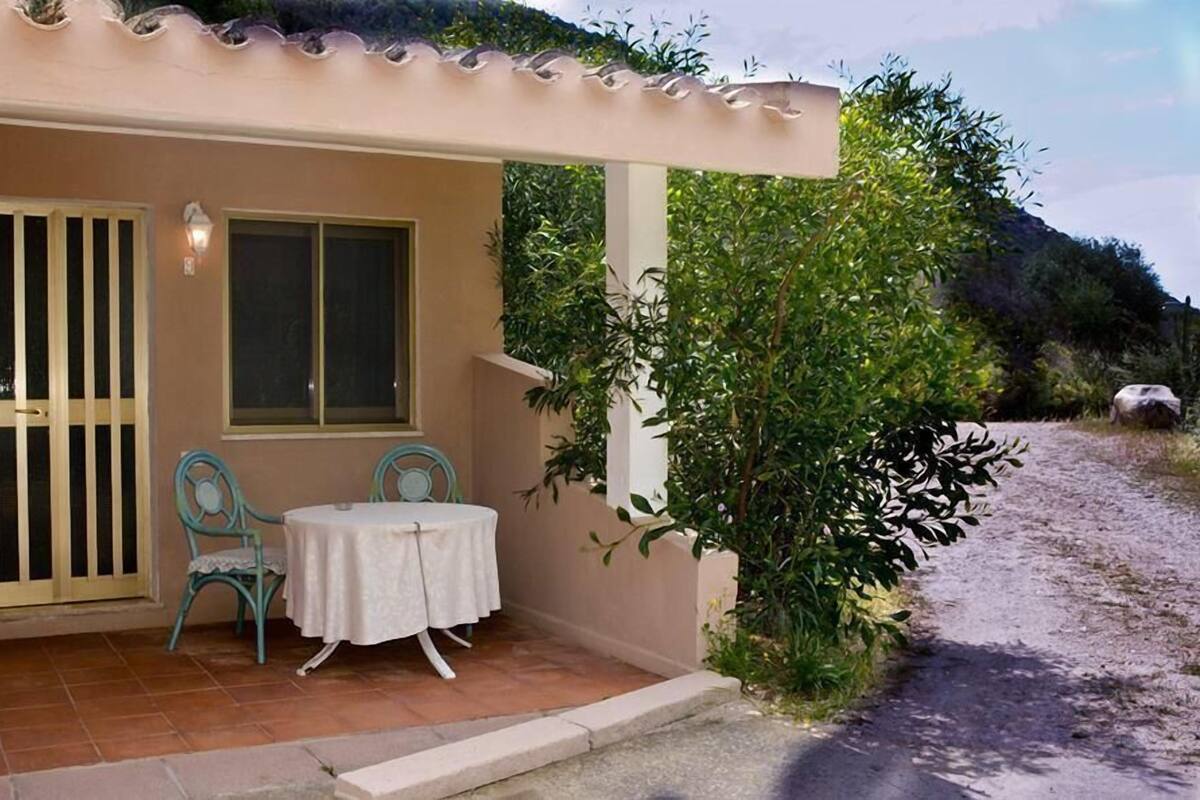 A covered outdoor seating area with a table and chairs, a window, and a door with a metal gate.