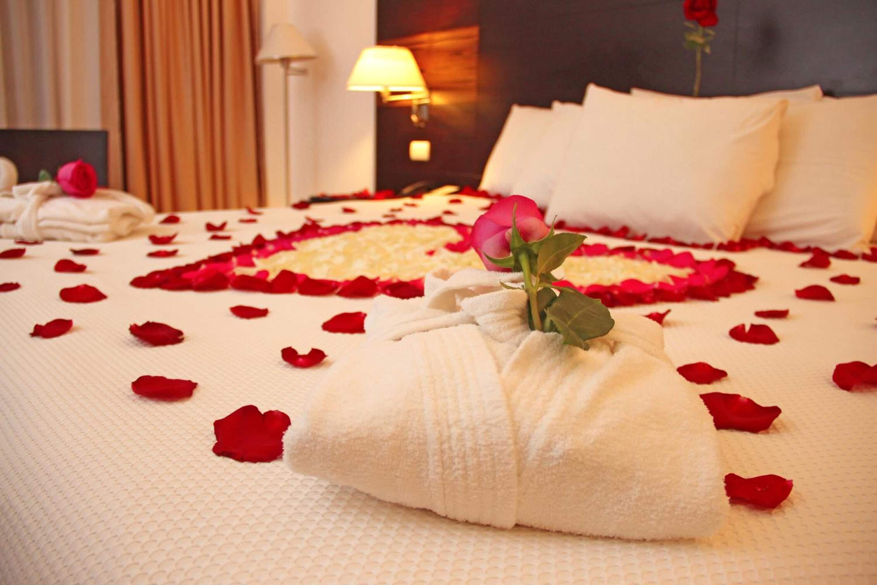 A neatly made bed with rose petals and a rose in a towel, a lamp, and a red rose on the wall.