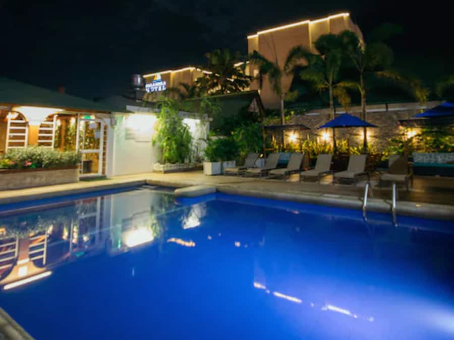 A swimming pool with lounge chairs and umbrellas, surrounded by greenery and a building with illuminated signage.
