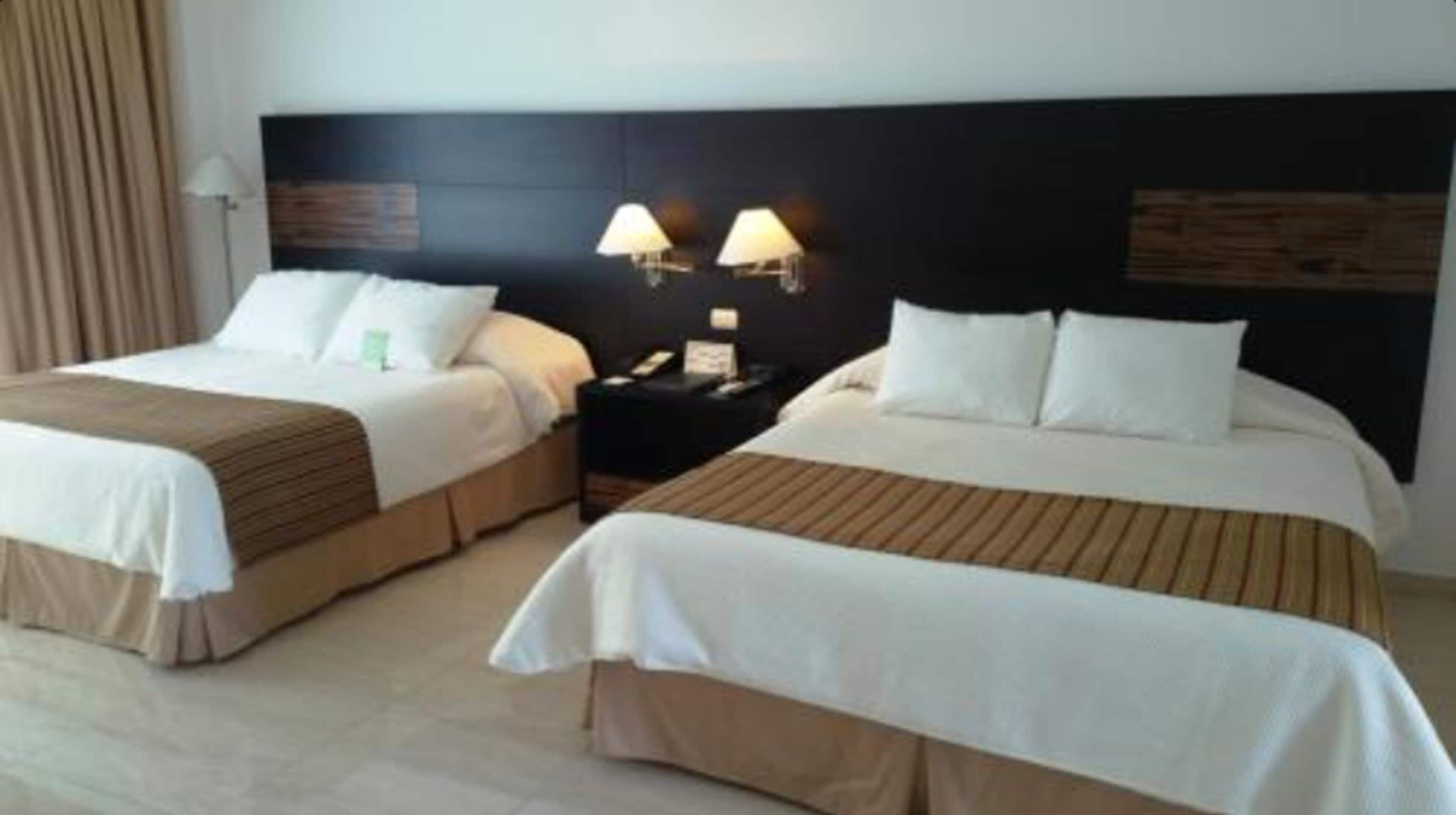 Habitación de hotel con dos camas, cada una con cabecero de madera y ropa de cama blanca. Hay dos mesitas de noche con lámparas.