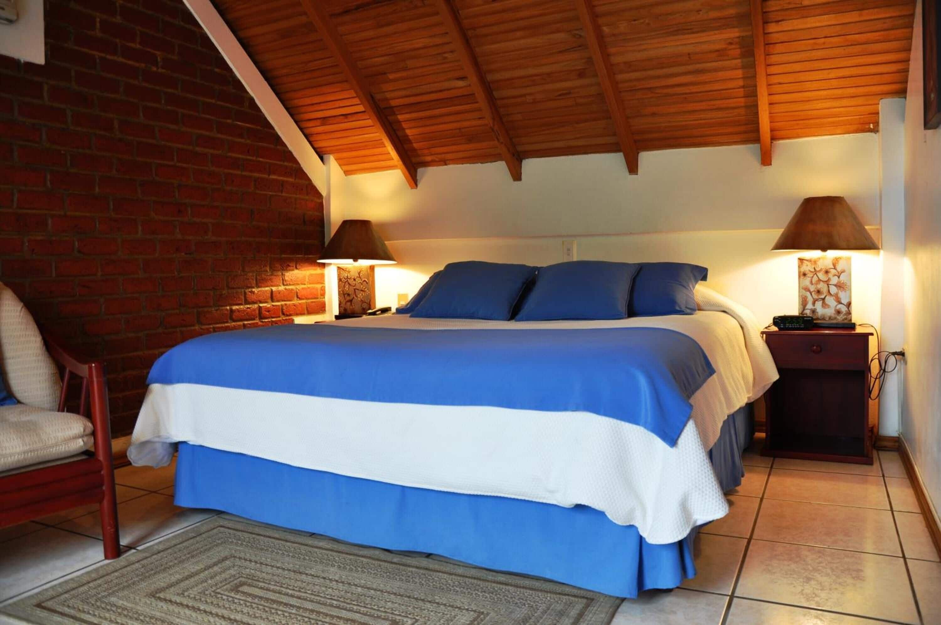 Una camera da letto con un letto grande, due lampade ai lati del letto, un muro in mattoni e un soffitto in legno.