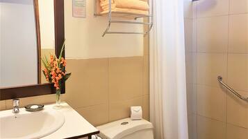 A bathroom with a sink, toilet, and shower. There is a towel rack with towels, a toilet paper holder, and a flower vase.