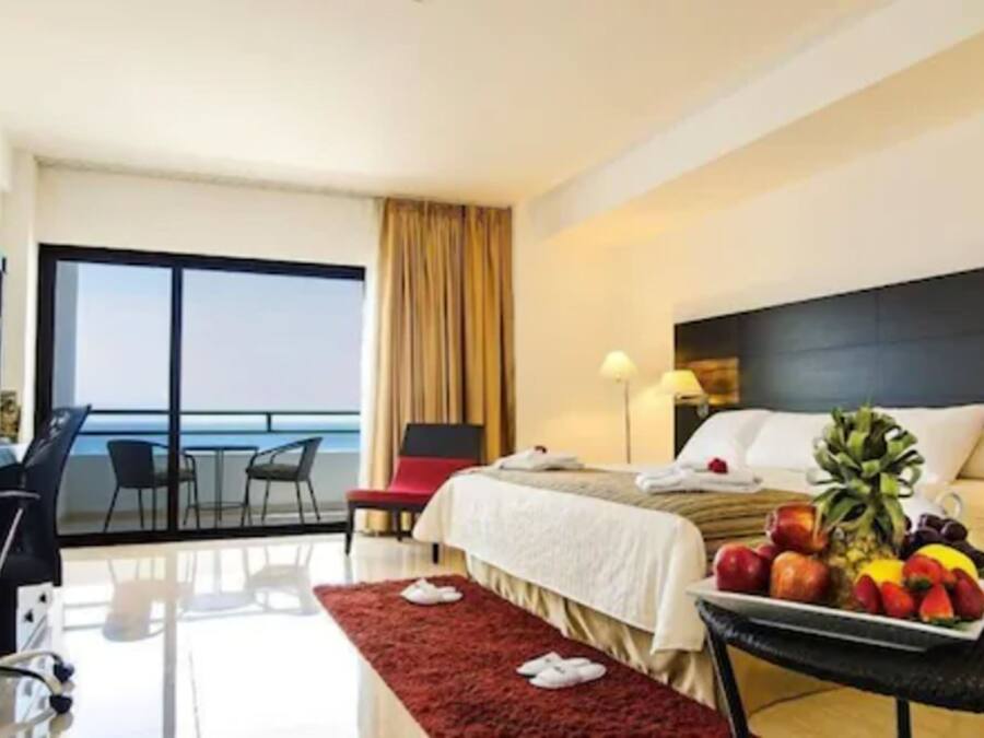 A hotel room with a bed, a dining table with fruit, a desk, and a view of the ocean.