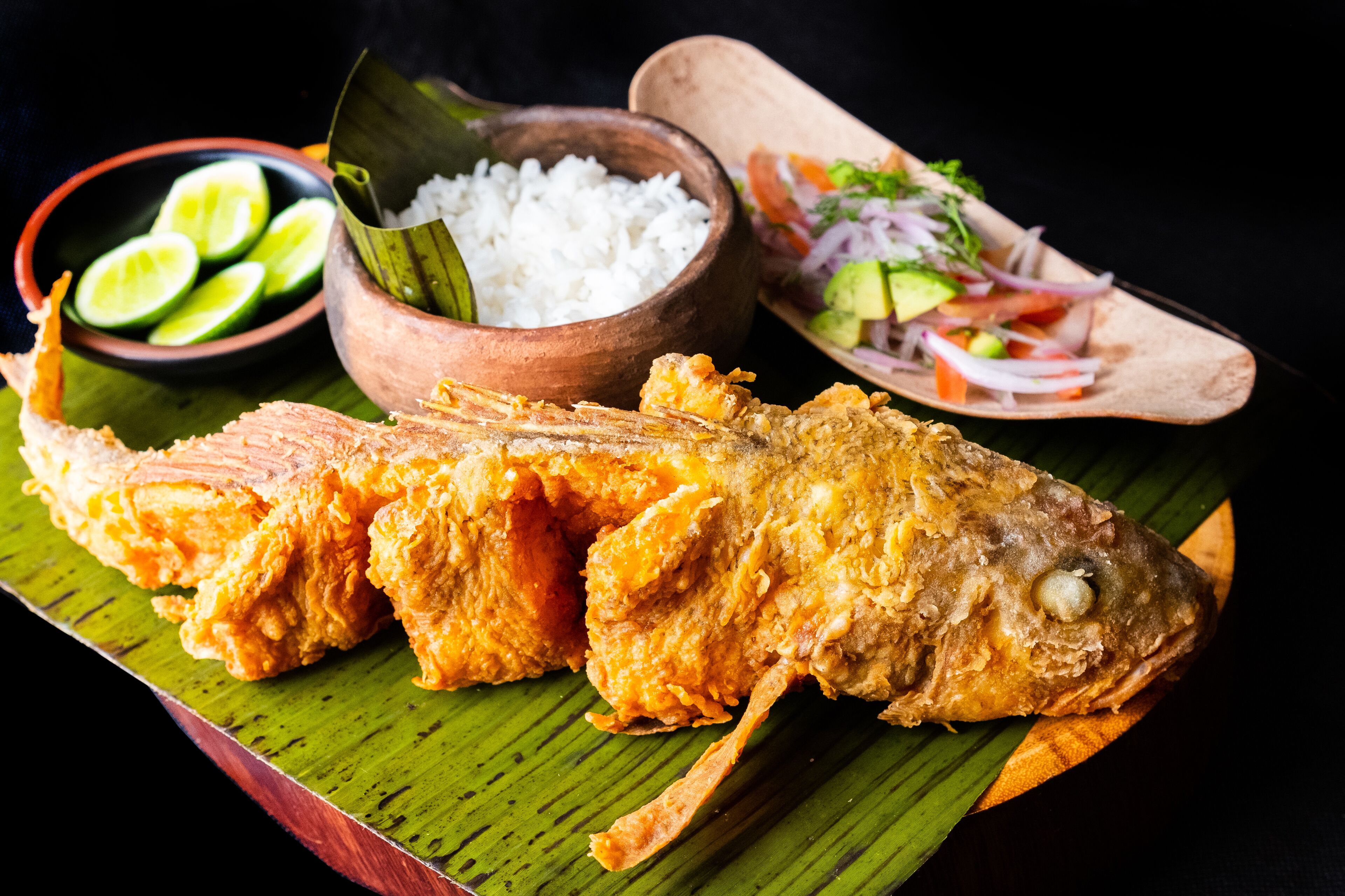 A whole fried fish on a banana leaf, accompanied by a bowl of white rice, a bowl of lime slices, and a wooden serving board with a salad.