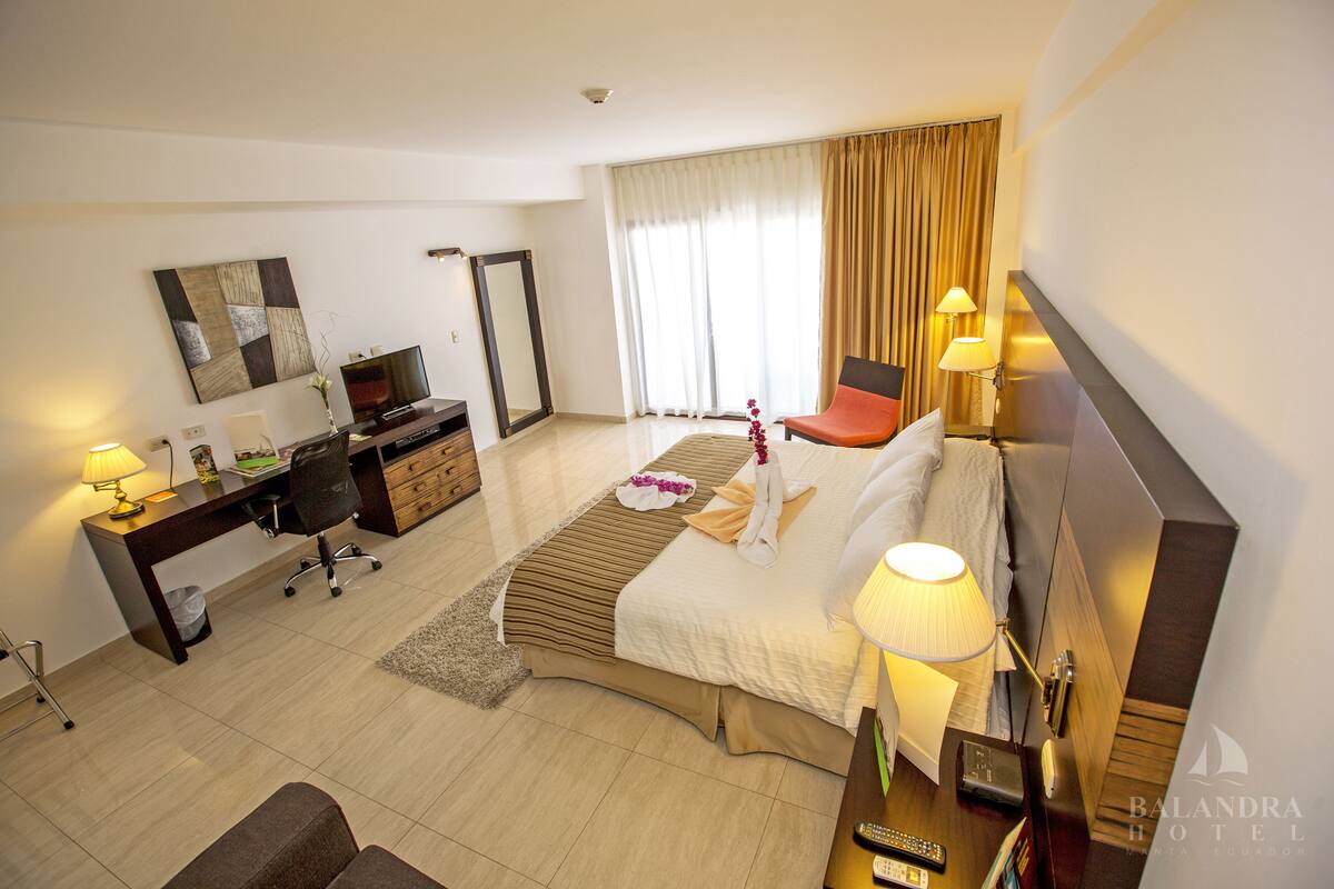 A hotel room with a bed, desk, chair, TV, and a large window with curtains.
