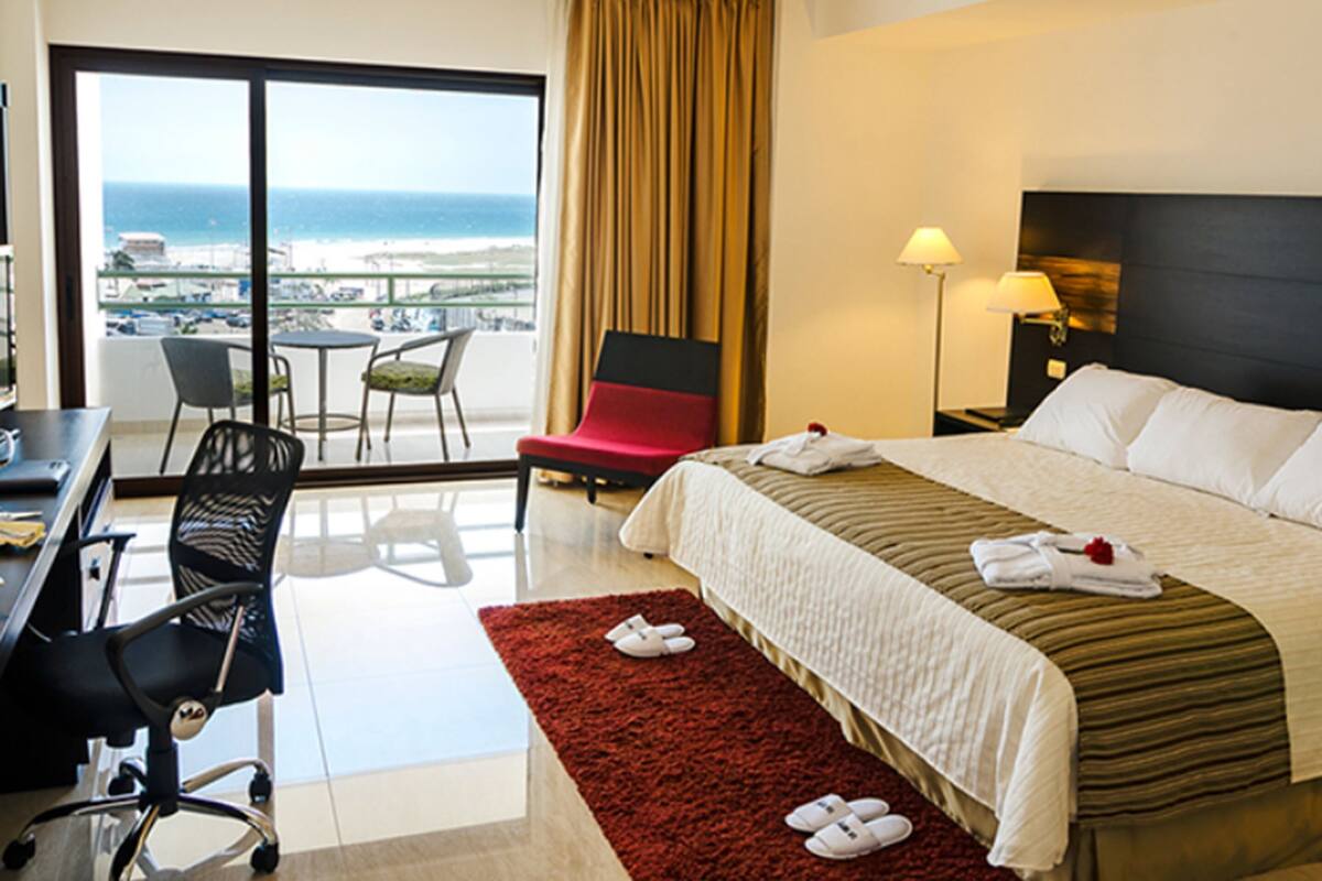 A hotel room with a bed, a desk, a chair, a lamp, and a view of the ocean.