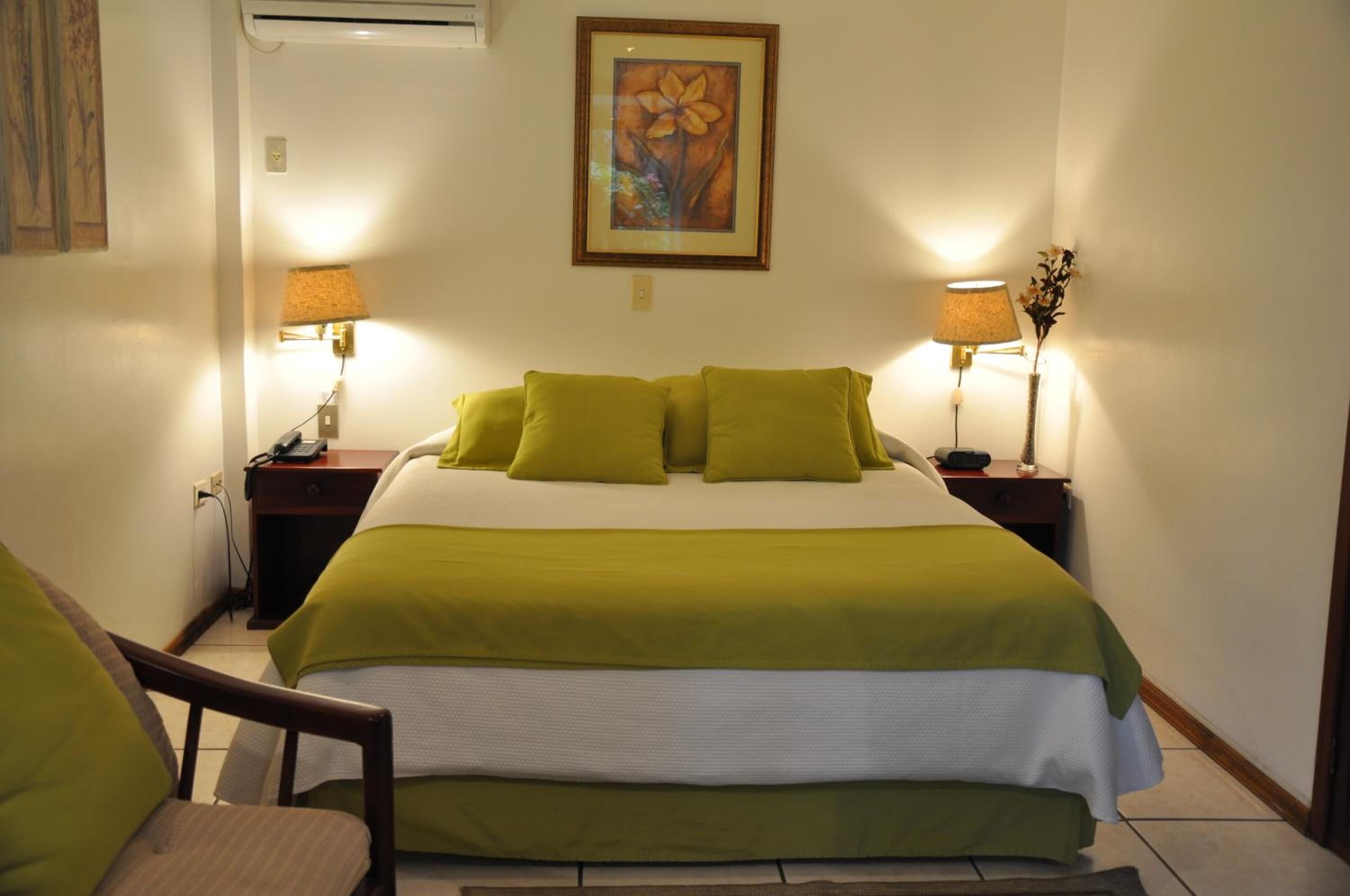 A neatly made bed with green bedding, two bedside lamps, a framed floral artwork on the wall, and a chair in the foreground.