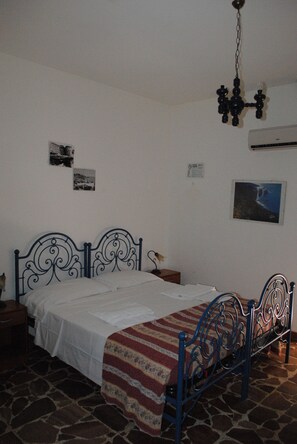 A bedroom with a metal bed frame, white bedding, a patterned throw blanket, a stone floor, a wall-mounted air conditioner, and a chandelier. - Calciufetta B&B (Alghero)
