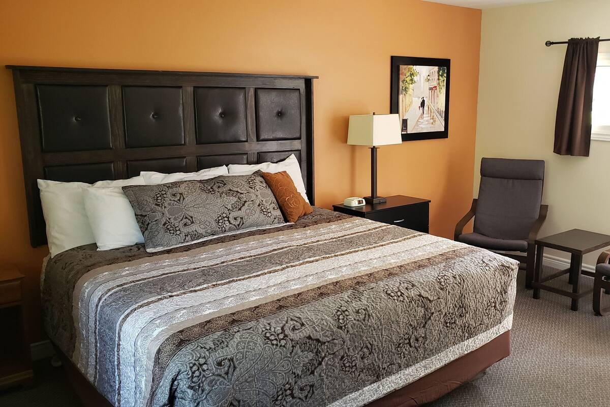 A hotel room with a large bed, a bedside table with a lamp, a chair, and a small table with a remote control.