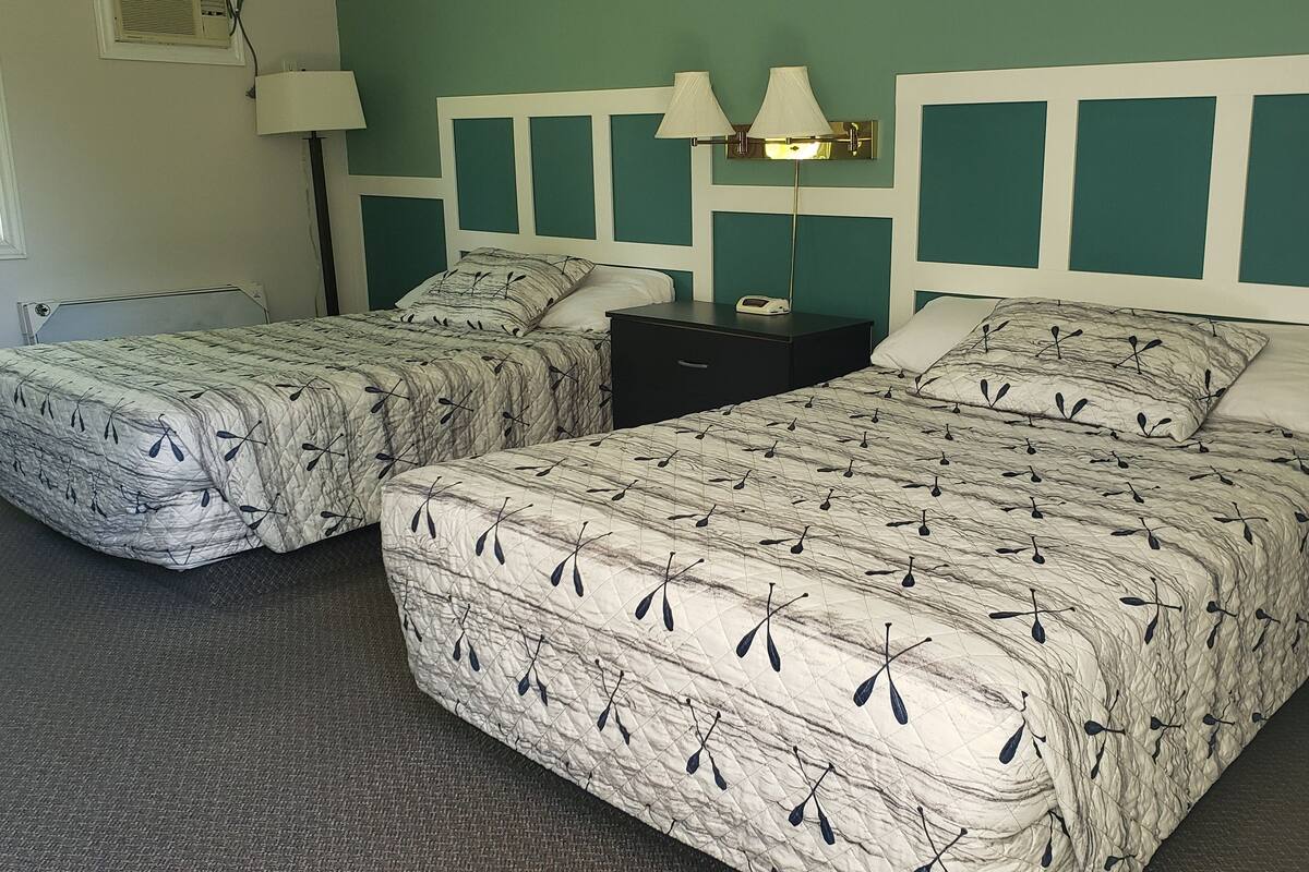 A room with two beds, each with a patterned bedspread and pillows, a bedside table with a lamp, and a wall-mounted air conditioning unit.