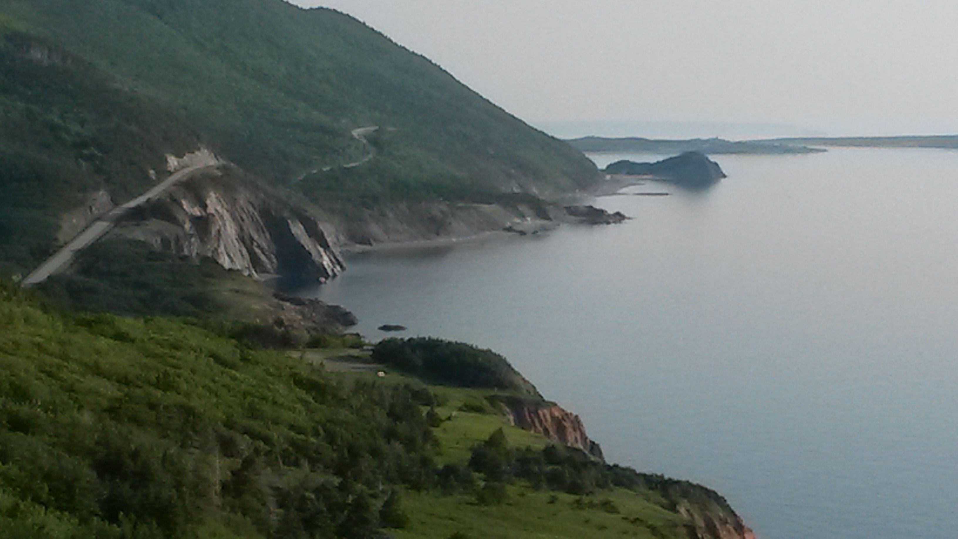 A coastal landscape with a winding road, green hills, and a calm sea.
