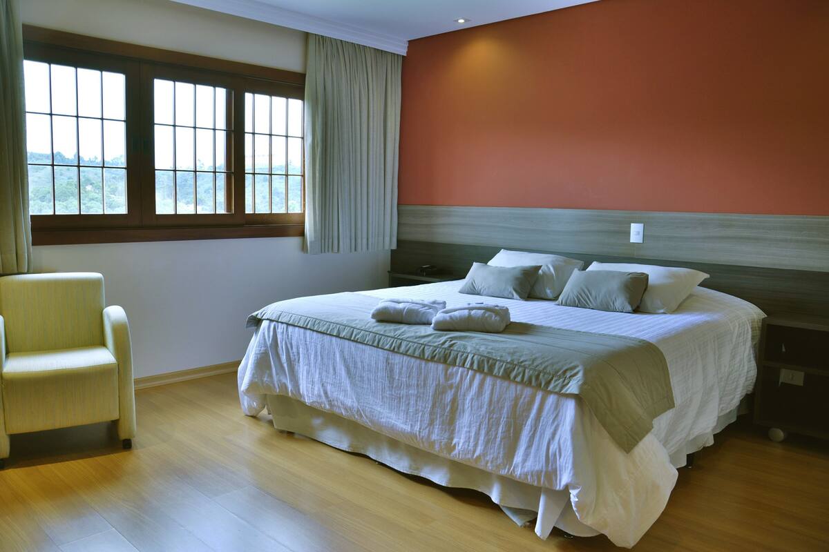 Un dormitorio con una cama grande, una silla, una ventana con cortinas y vistas al verdor exterior.