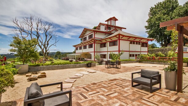 Un amplio patio exterior con suelo de baldosas estampadas, una zona de estar con dos sillas y un edificio grande de esquema rojo y blanco al fondo.