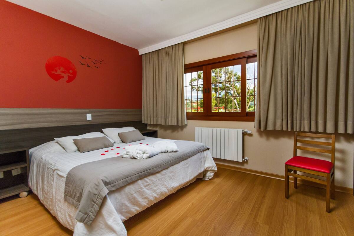 Un dormitorio con cama, ventana con cortinas, una silla de madera y pared roja con un diseño circular.