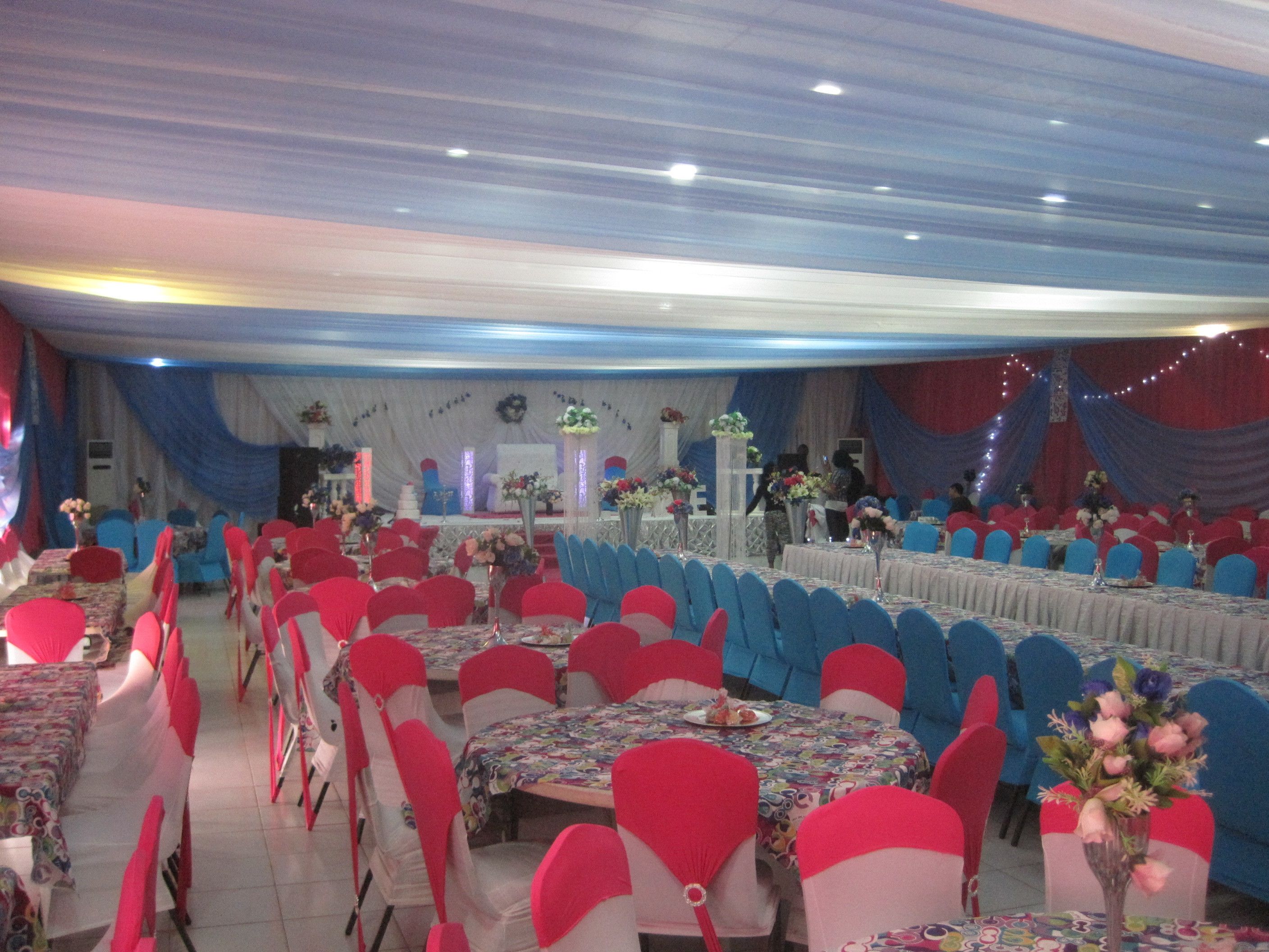 Un salón de banquetes con filas de mesas y sillas, un escenario con un telón de fondo y un techo decorado con telas colgantes.