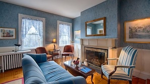 A living room with a blue sofa, striped armchair, fireplace, and large windows with curtains. - Sherburne Inn (Nantucket)