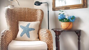 A wicker armchair with a star-shaped pillow, a small table with a wicker basket holding blue flowers, and a mirror with a gold frame on the wall. - Sherburne Inn (Nantucket)