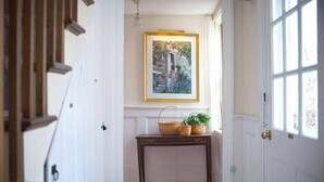 A hallway with a framed painting, a wooden console table with baskets, and a door with a brass handle. - Sherburne Inn (Nantucket)