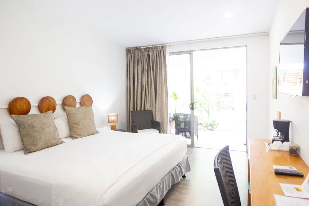 Ein Hotelzimmer mit einem großen Bett, einem Schreibtisch und einem Sessel. Es gibt ein Fenster mit Vorhängen, eine Kaffeemaschine und eine Pflanze.