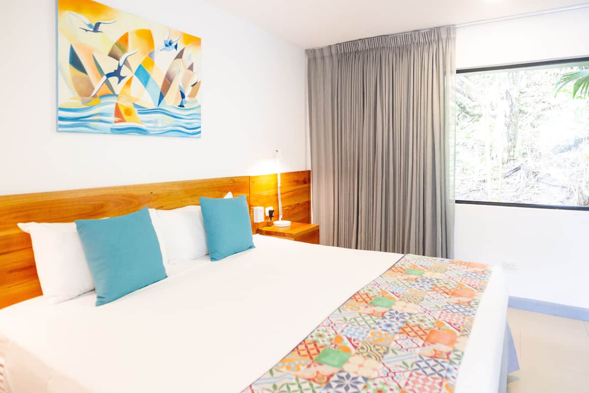 Ein Hotelzimmer mit einem großen Bett, einem Holzkopfteil, einer farbenfrohen Tagesdecke, einem Nachttisch mit Lampe, einem Fenster mit Vorhängen und einem Wandbild.