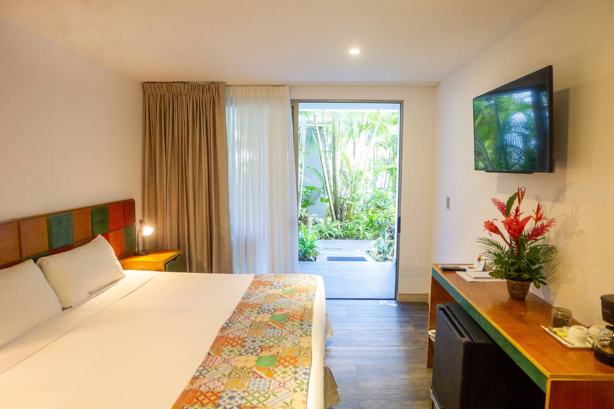 Ein Hotelzimmer mit Bett, Schreibtisch, Fernseher und Blick auf einen Garten durch die Tür.