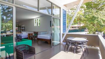 A modern hotel room with a glass wall, a balcony with a table and chairs, and a bed with a green chair and a small table.