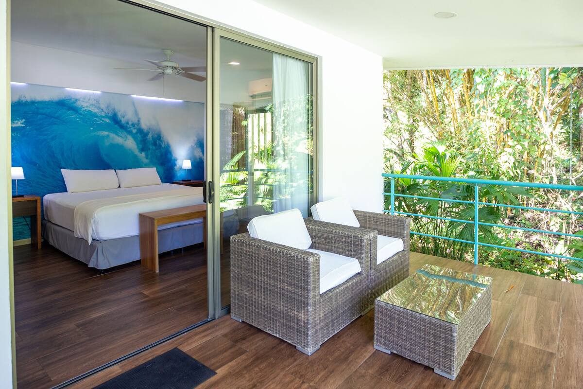 Ein Schlafzimmer mit Bett, Sofa und einem kleinen Tisch. Eine Glastür führt zu einem Balkon mit Pflanzen.