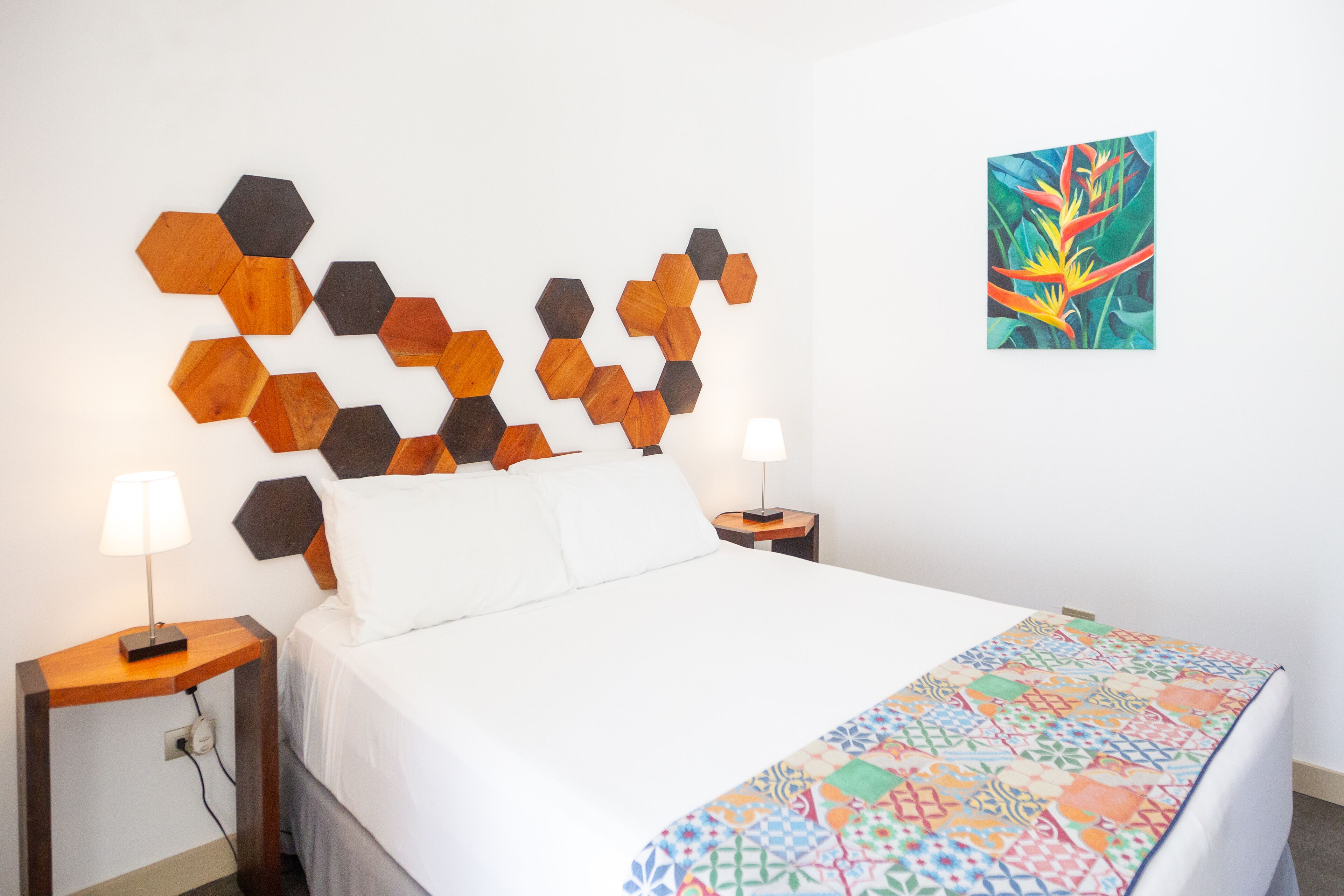 A hotel room with a bed, a wooden hexagonal headboard, a colorful quilt, and a painting on the wall.