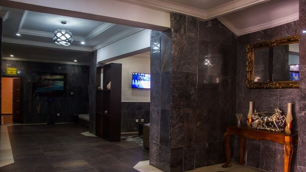 A lobby with dark marble flooring, a wooden console table with decorative items, a large mirror with an ornate frame, and a television mounted on the wall. - Eastgate Hotel Lekki (Lekki)