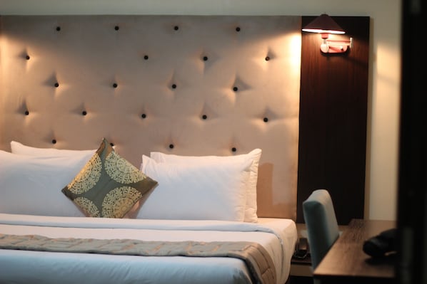 A hotel room with a tufted headboard, a bed with white linens, a decorative pillow, a nightstand, and a wall-mounted lamp.
