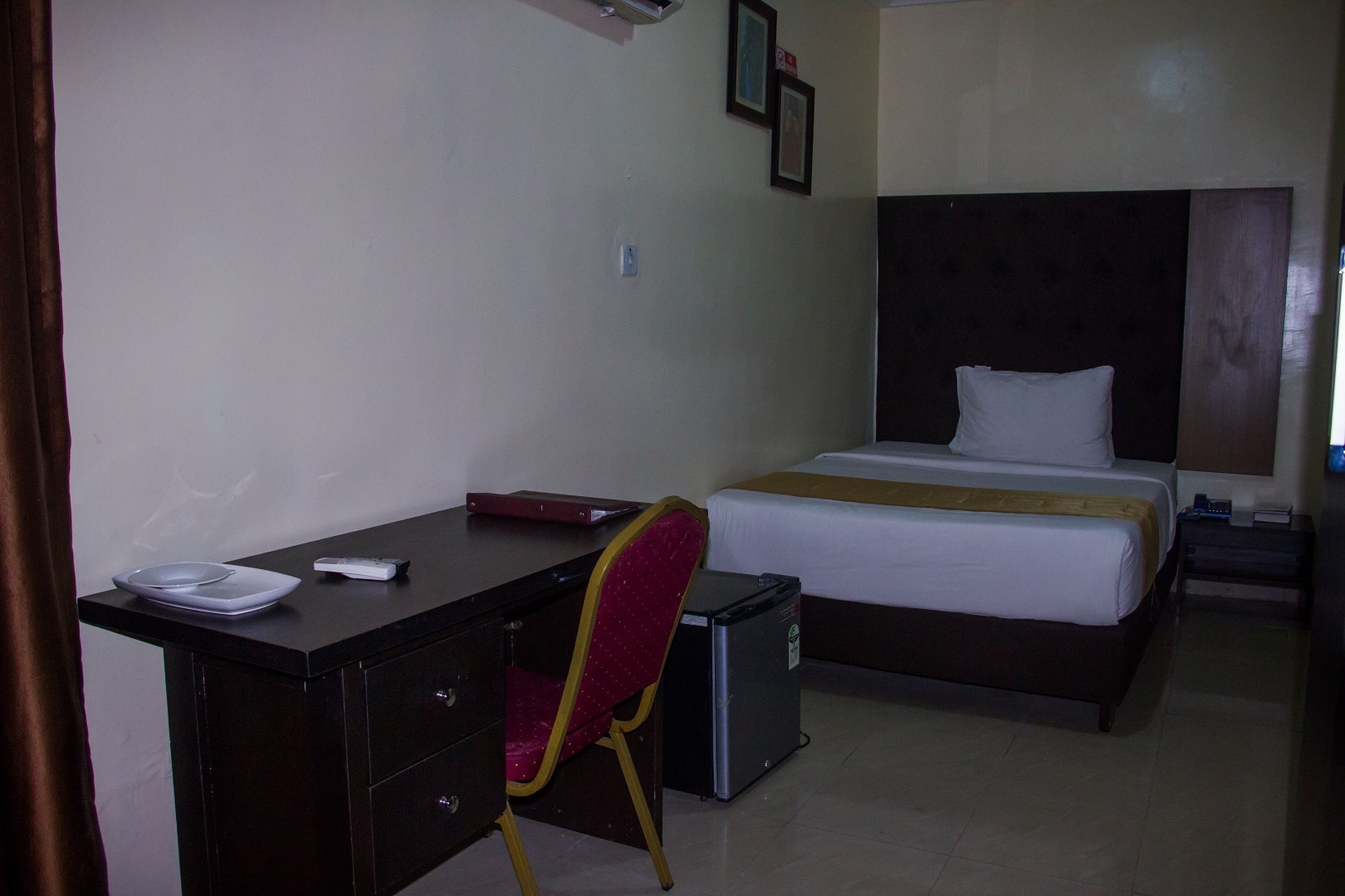 A hotel room with a bed, a desk with a chair, a small refrigerator, and a bedside table with a lamp.