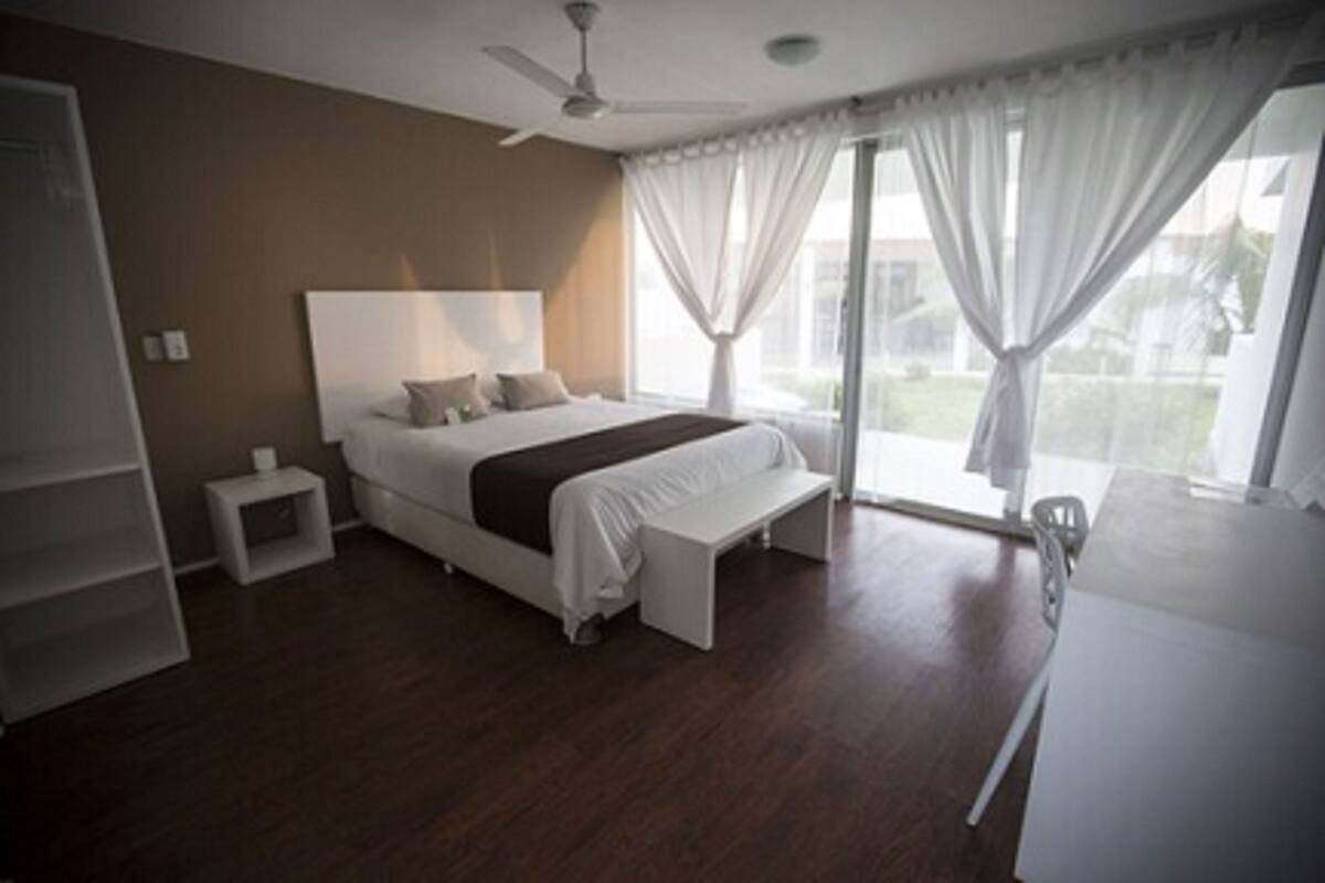 Un dormitorio moderno con una cama grande, una mesita de noche, una lámpara pequeña y vistas al exterior a través de un amplio ventanal con cortinas.