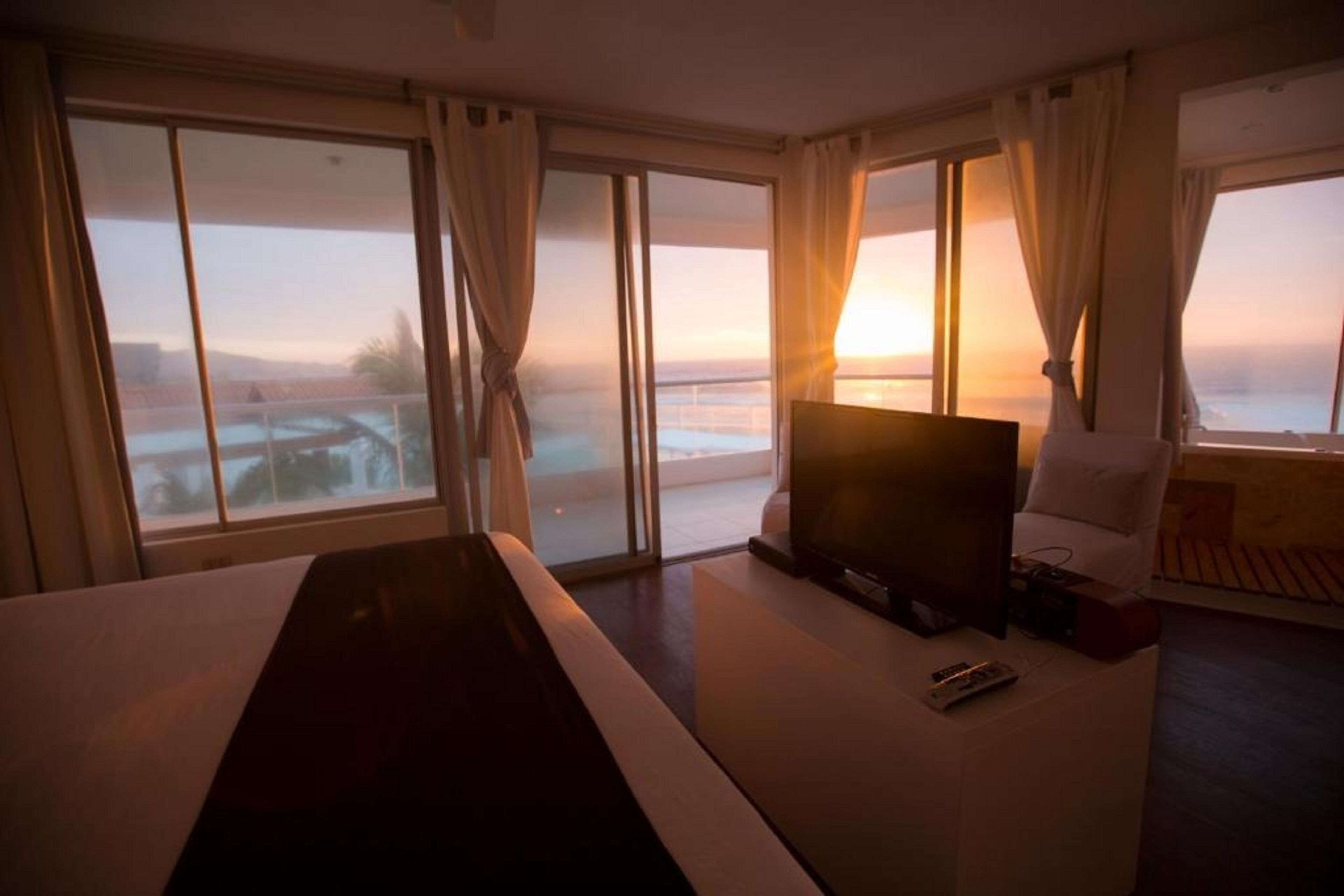 A hotel room with a large bed, a TV, and a sofa. There are large windows with curtains, and a view of a body of water and a bridge.