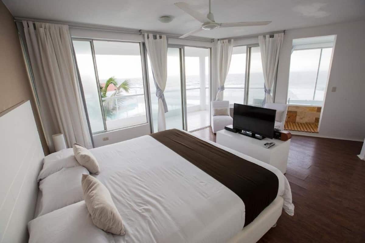 Habitación de hotel con una cama grande, un televisor en una mesita auxiliar y un balcón con vista al océano.