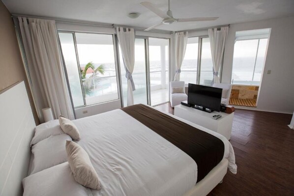 A hotel room with a large bed, a TV on a side table, and a balcony with a view of the ocean. - Suites del Mar (Máncora)