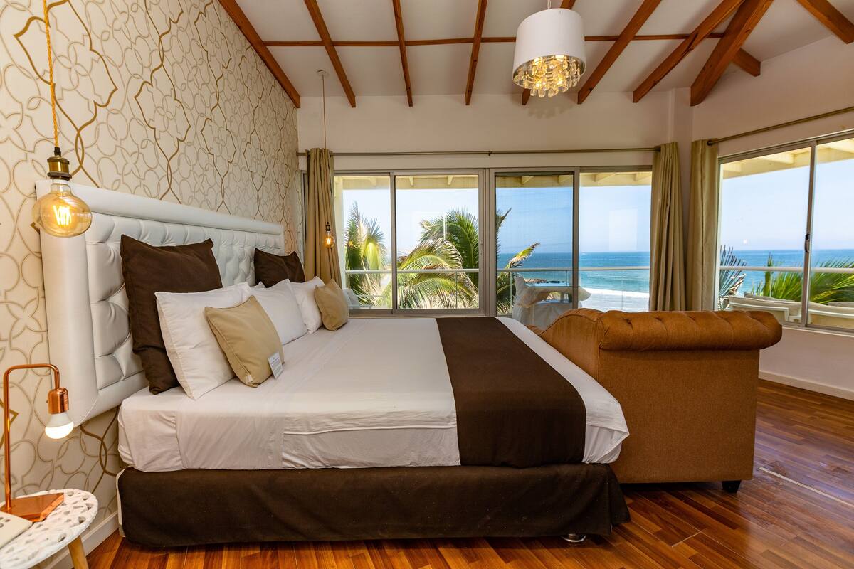 Un dormitorio amplio con una cama grande, un sofá y vista al océano a través de amplias puertas correderas.
