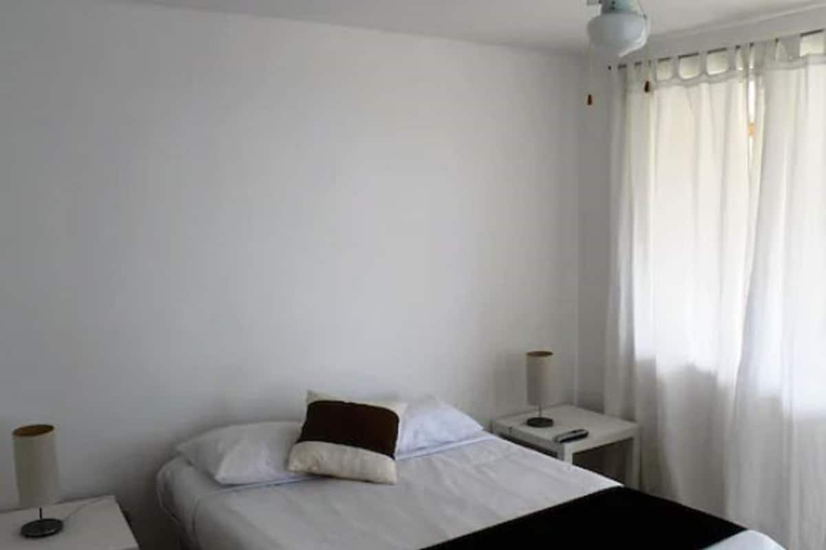 Una habitación sencilla y luminosa con cama, dos mesitas de noche y una lámpara.
