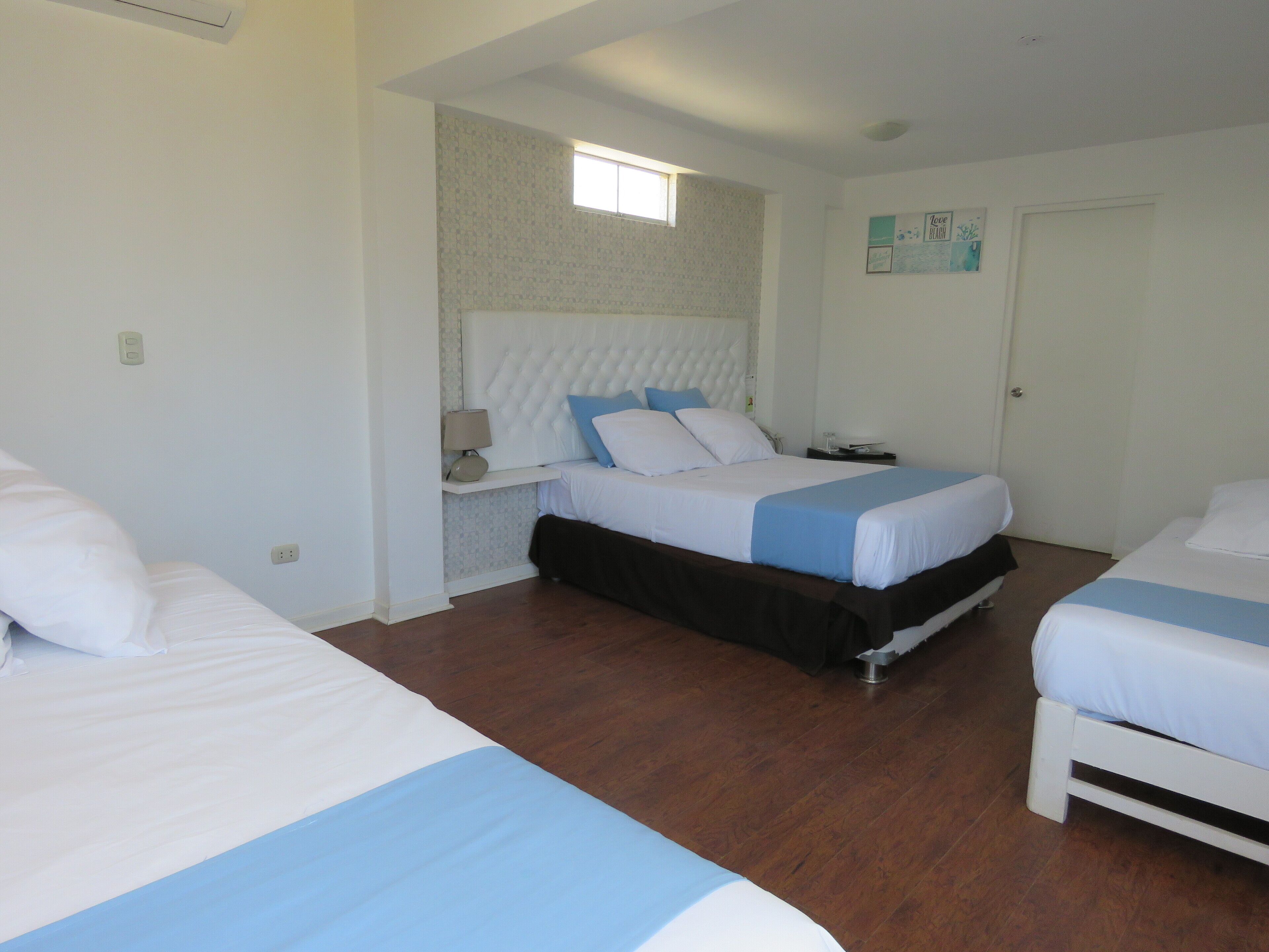 A hotel room with two beds, a small bedside table with a lamp, a wall-mounted shelf, and a door.