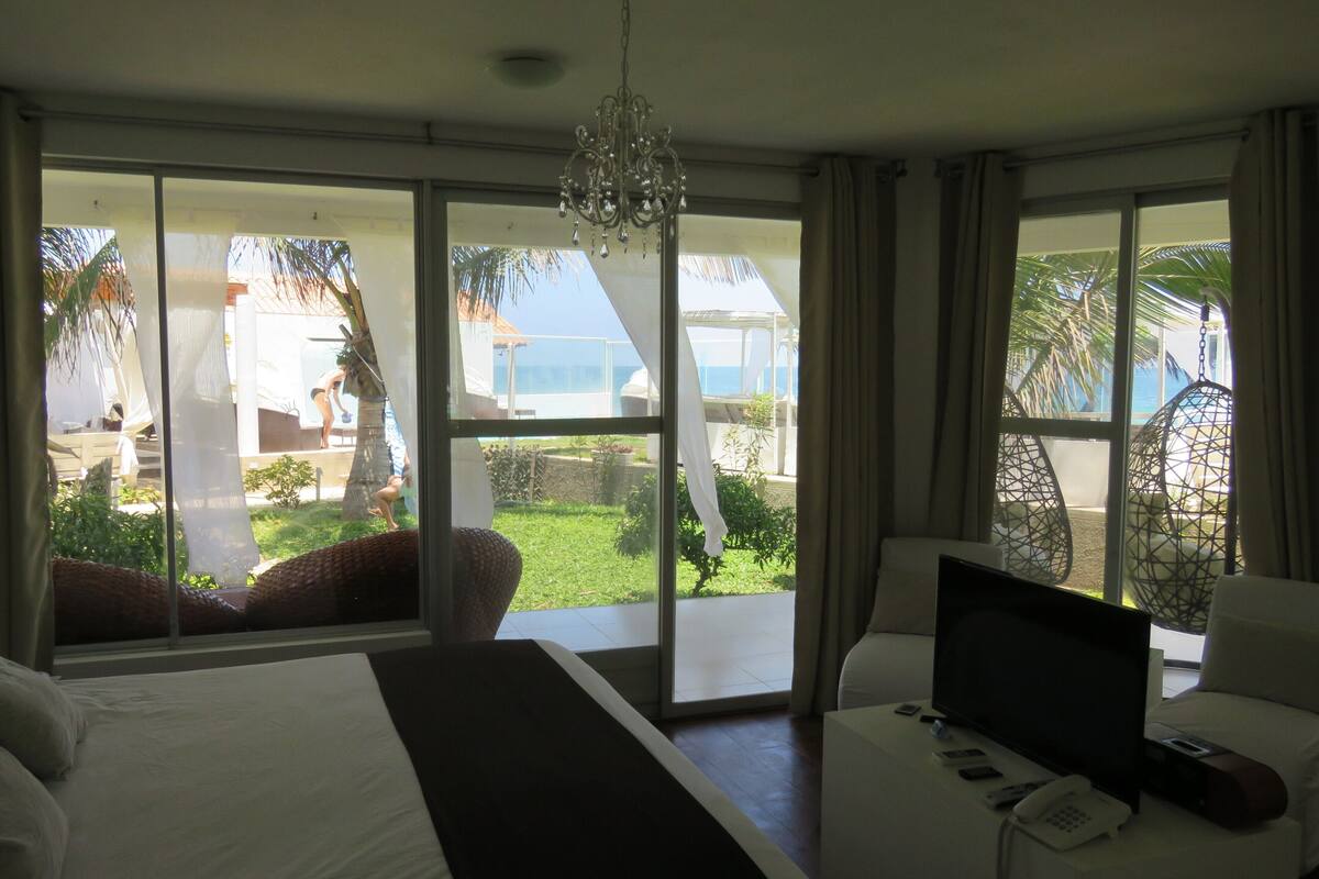 Habitación de hotel con una cama grande, televisión y teléfono sobre una mesita de noche. Hay una puerta corrediza de vidrio que da a un balcón con vistas a palmeras y una piscina.