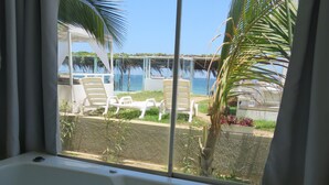 Un baño con bañera, una ventana con cortinas y vistas a una zona de piscina con sillas de descanso y palmeras.