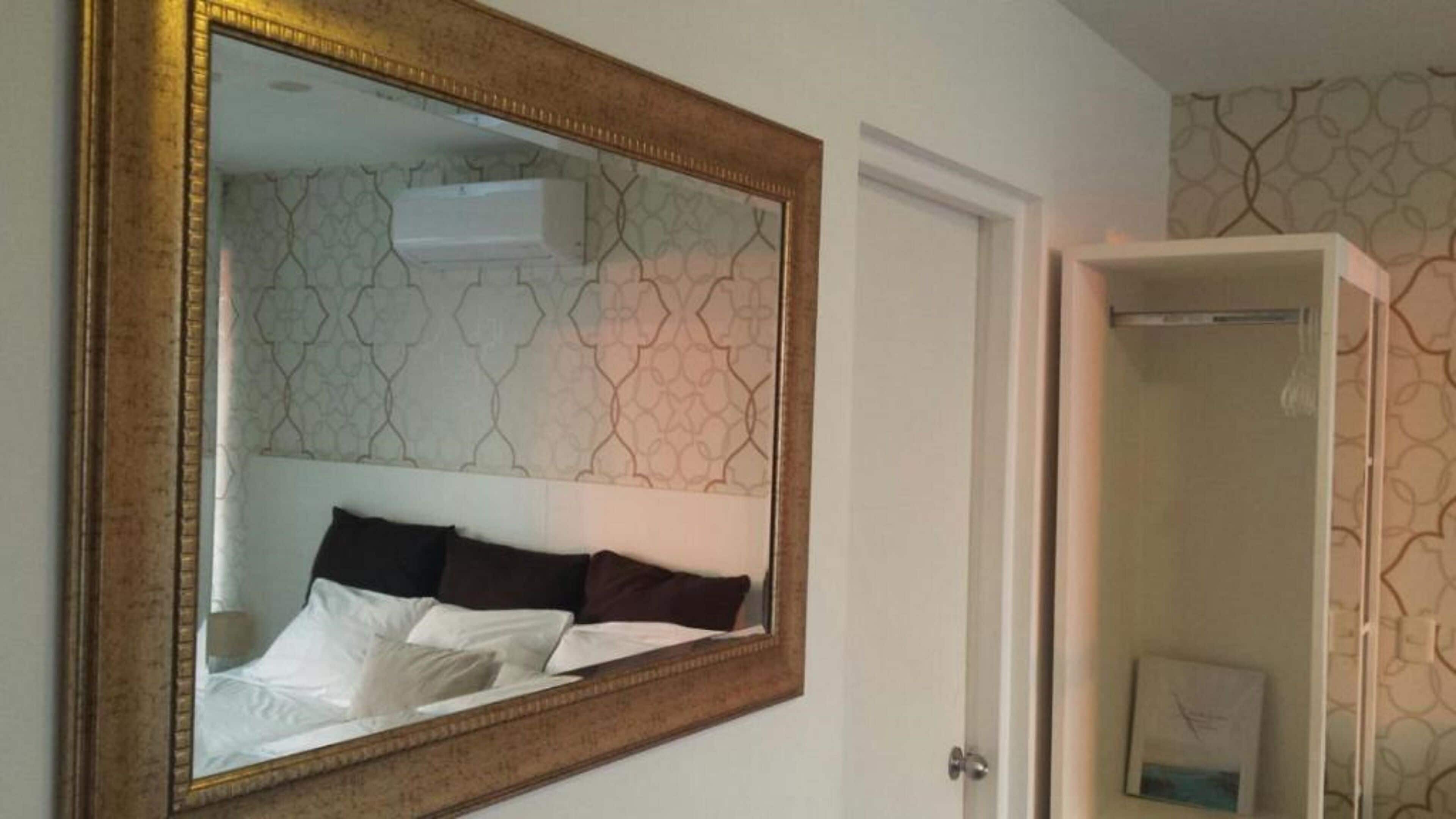 A bedroom with a large mirror, a bed with white and dark pillows, and a patterned wallpaper.