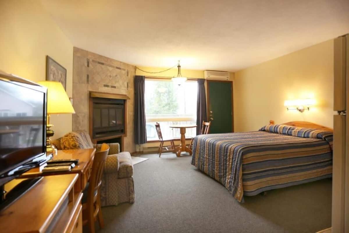 A hotel room with a bed, a desk with a television, a fireplace, a dining table with chairs, and a window with curtains.